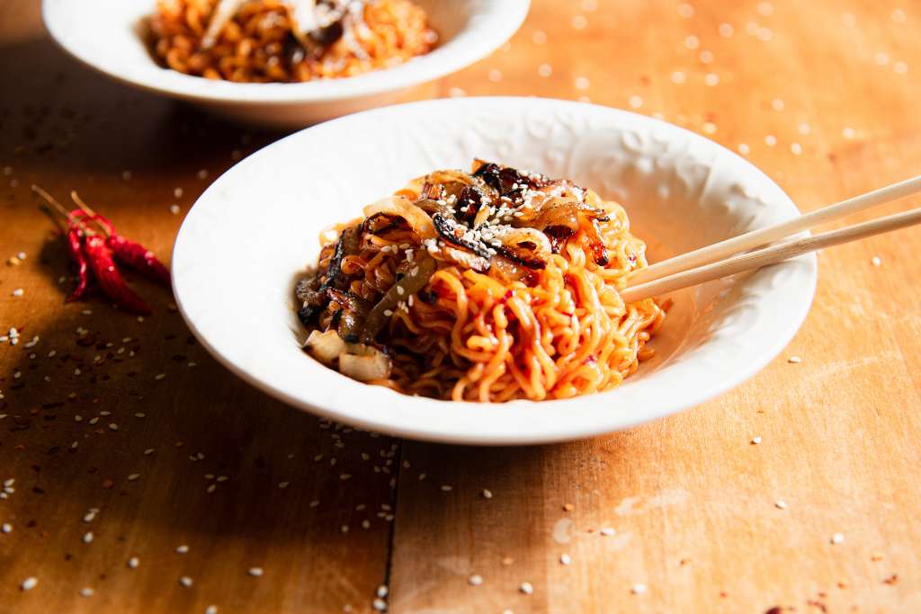 a bowl of noodles with chopsticks stuck in it, there are some dried peppers next to the bowl and part of another bowl in the back of the photo