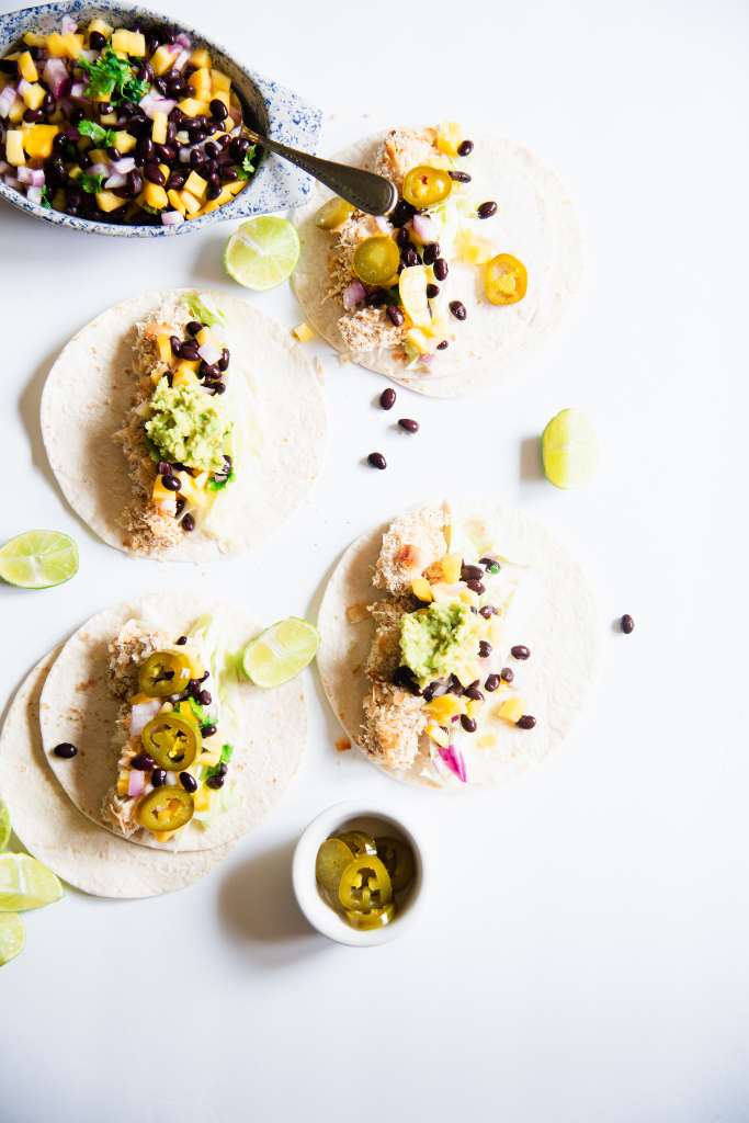 four fish tacos laying open, there is a dish of mango salsa at the top on the photo and a dish of jalapenos at the bottom. There are some black beans and lime wedges, around the tacos