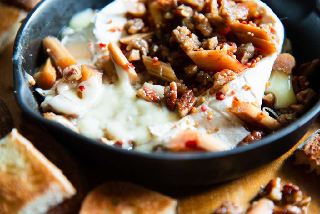 a close up of melted brie in a cast iron skillet, the brie is topped with rhubarb and pecans, you can part of a few pieces of toast in front of the pan 