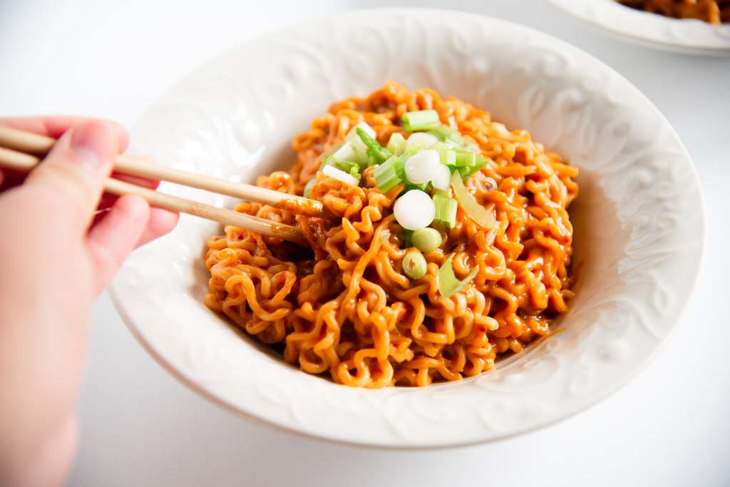 a bowl of spicy peanut ramen with sliced green onions on top, someone is starting to pick up a few noodles with a pair of chopsticks 