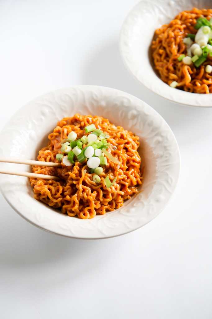 a bowl on noodles with green onions on top, and chop sticks stuck in them. You can see part of another bowl of noodles behind it 