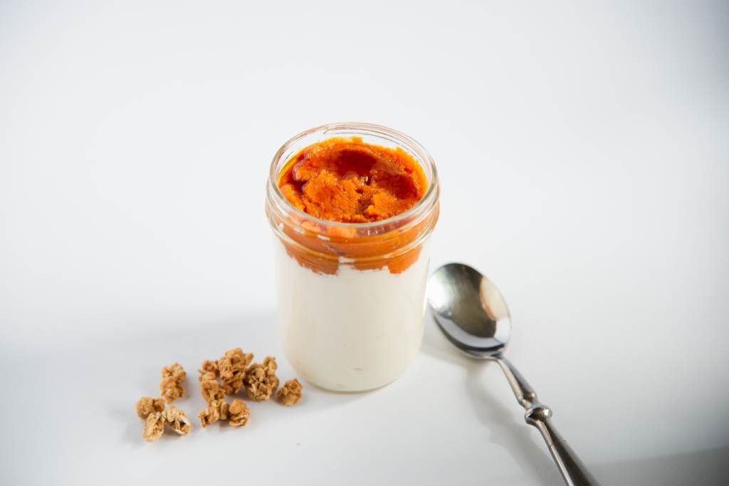 a jar of yogurt with pumpkin and maple syrup in it, there is a spoon and some granola next to it 
