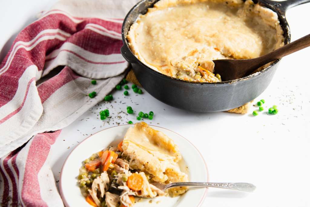 an overhead picture of a pan of pot pie and a plate with chicken pot pie and a fork on it. There is also a tea towel and some peas by the pan and plate