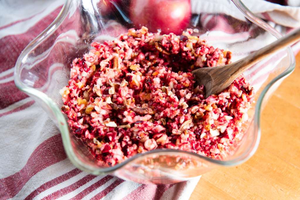 a glass bowl of cranberry relish with a wooden spoon in it, the bowl is set on a towel and your can see a couple f apples behind  the bowl