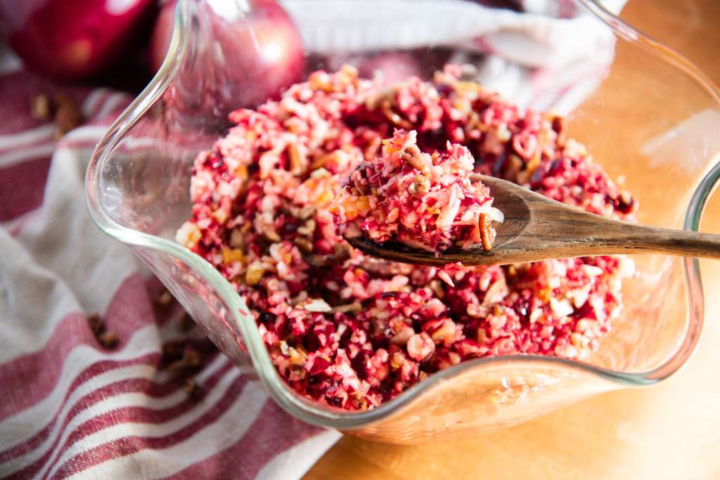 a spoonful of cranberry relish being held over a bowl of it, the bowl is sitting partly on a tea towel and there are some apples and pecans by it