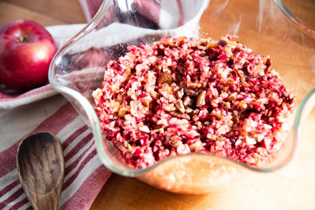 a close up of a glass bowl of cranberry relish, there is a red and white tea towel, an apple and a wooden spoon next to the bowl 