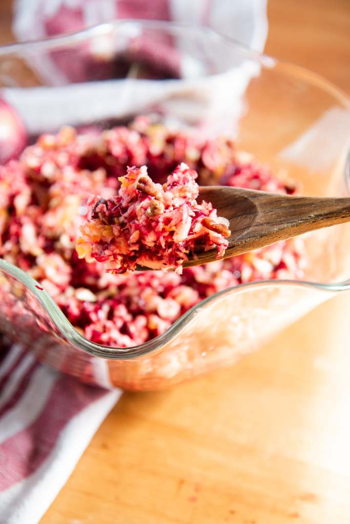 a close up of a spoon of cranberry relish being held over the bowl of relish, you can see part of the red and white tea towel behind the bowl 