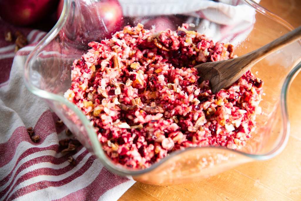 a bowl of cranberry relish with a wooden spoon in it. There is a red and white tea towel, a few apples, and some chopped pecans next to the bowl