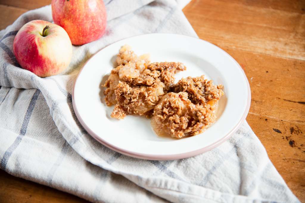 a plate of apple crisp set on a gray and white tea towel. There are two apples sitting next to the plate 