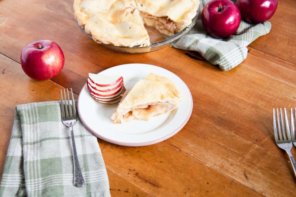 a pie of pie on a plate with slices of apple, there is a tea towel with a fork on it next to the plate, and some whole apples, more forks and the rest of the pie around the plate 