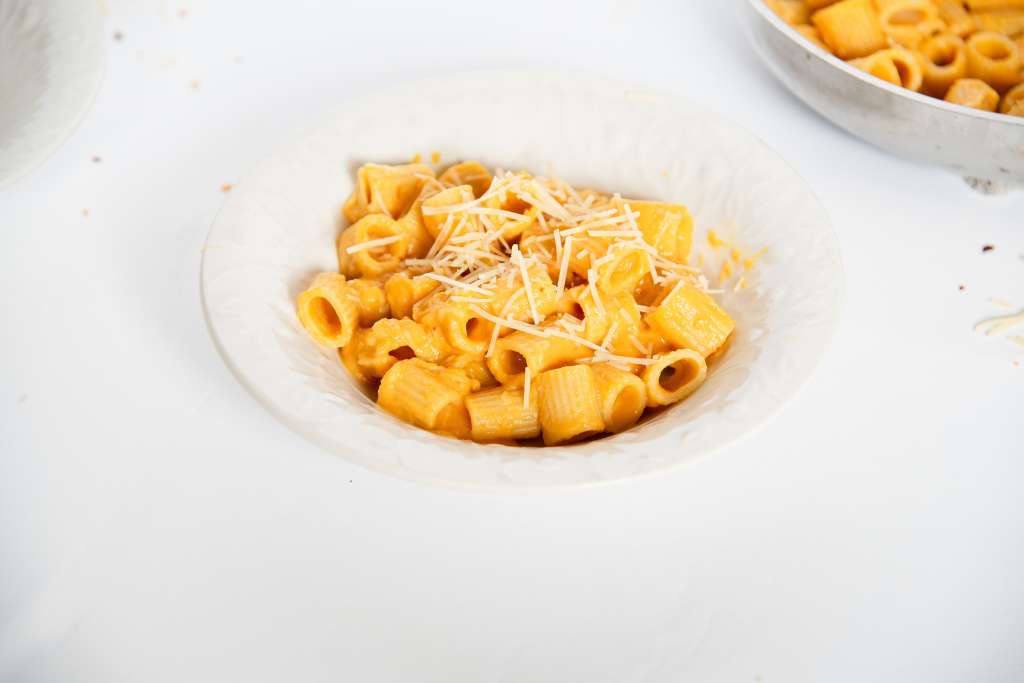 a close up of a bowl of pasta with carrot sauce and shredded cheese on top, you can part of another bowl and the pan of pasta in the background