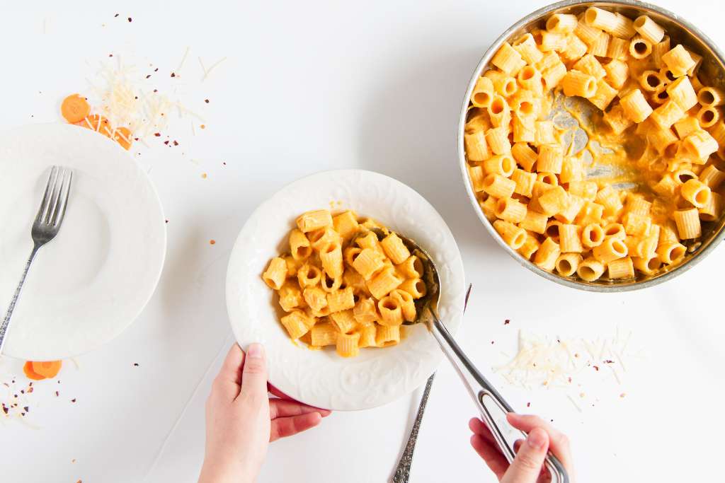 someone scooping a spoonful of sauce covered noddles into a bowl, there is a fork and the pan of noodles to one side, and an empty bowl with a fork in it on the other side 