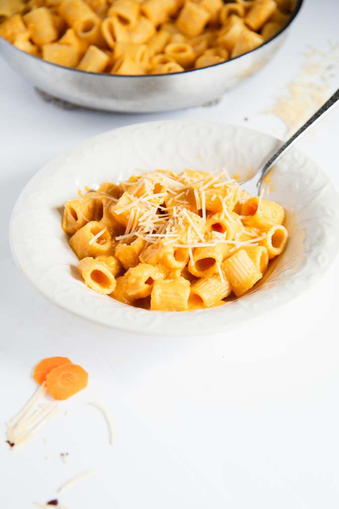 a bowl of pasta that has been mixed in carrot sauce, there is a fork in the bowl and shredded cheese on top of the noddles. The pan of the noodles in behind the bowl, and there are a few carrot slices and some cheese in front of the bowl