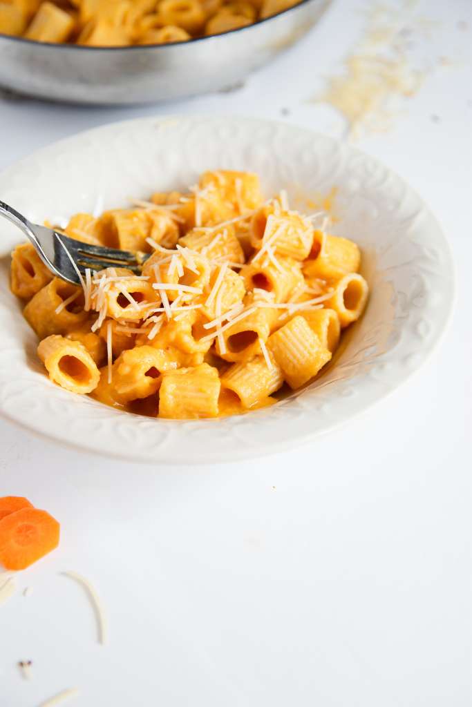 a fork full of noddles being held just above a bowl of noddles covered in sauce and shredded cheese, you can see part of the pan of noddles and a few carrot slices next to the bowl