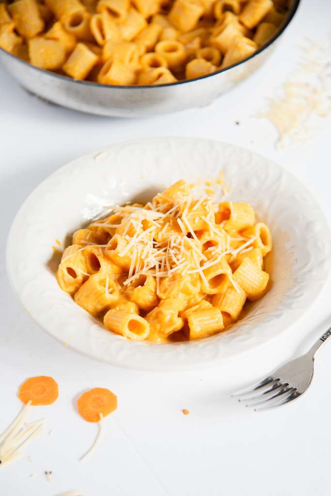 a bowl of noddles coated in carrot pasta sauce with shredded cheese on top. There is a fork next to the bowl and you can see part of the pan of noodles in the background 