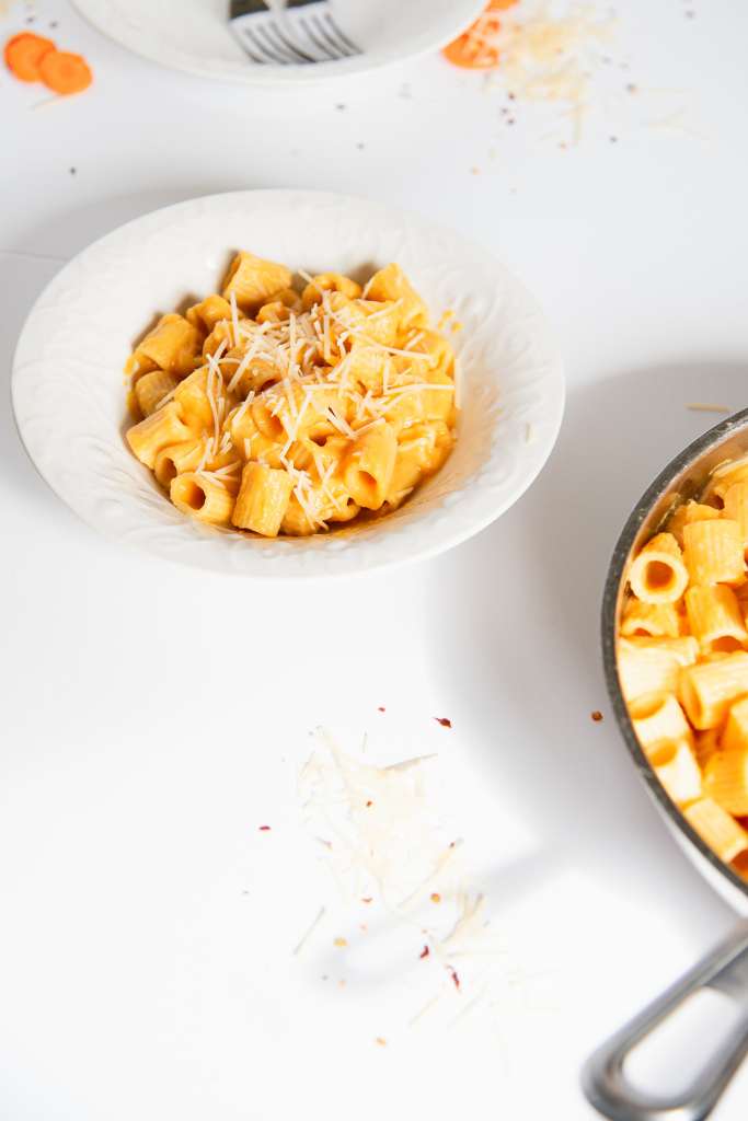 a bowl of carrot sauce covered noodles with cheese on top, you can see part of the pan to the side of the bowl and an empty bowl with two forks in it, and some cheese and carrot slices behind the bowl