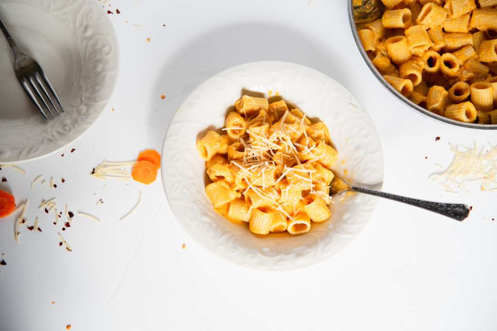 a bowl of noodles covered in sauce and shredded cheese with a fork stuck in it. There is a pan of noodles to one side and an empty bowl with a fork in it to the other