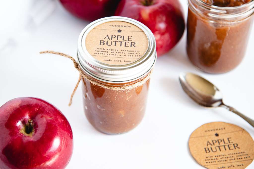 a jar of apple butter with a piece of string tired around it and a label on it. There is uncovered jar behind it, and some apples, a spoon and another label around it 