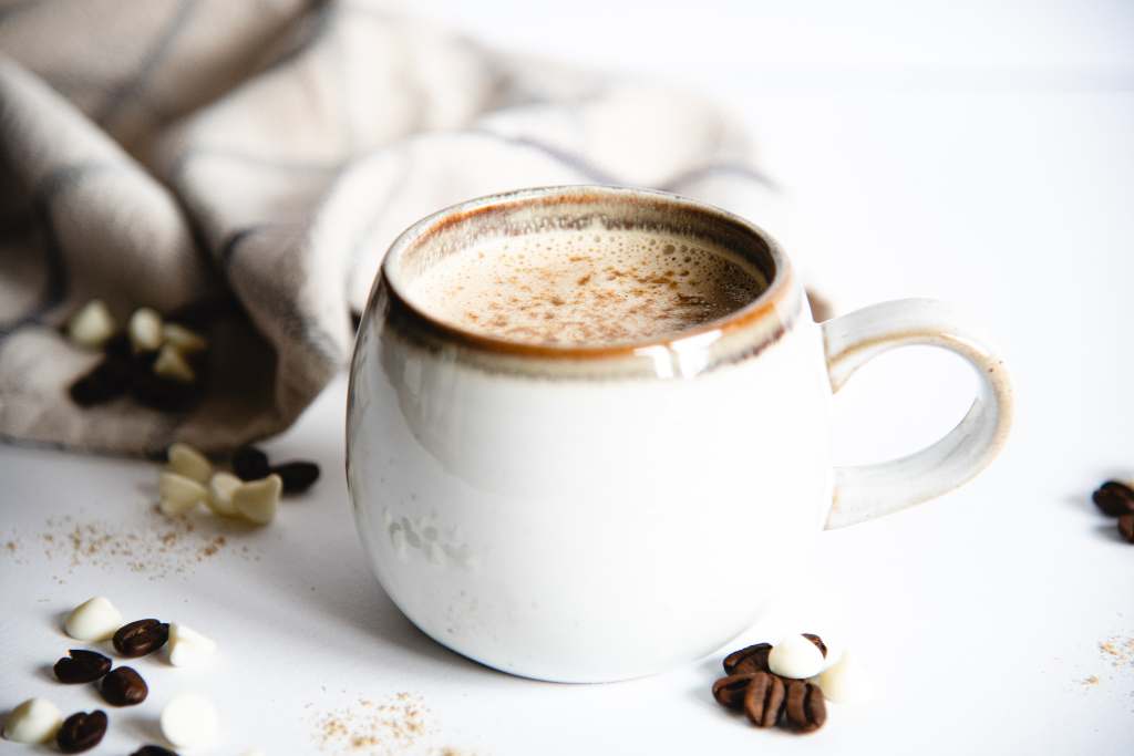 a close up of a mug of white chocolate espresso, there are some coffee beans, white chocolate chips, and cardamom sprinkled around it, and you can see part of a tea towel behind the mug