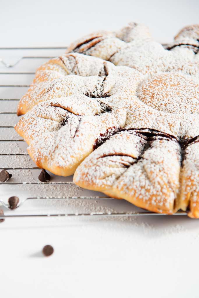 a cut off picture of a finished star bread topped with powder sugar, on a cooling rack with a few chocolate chips next to it