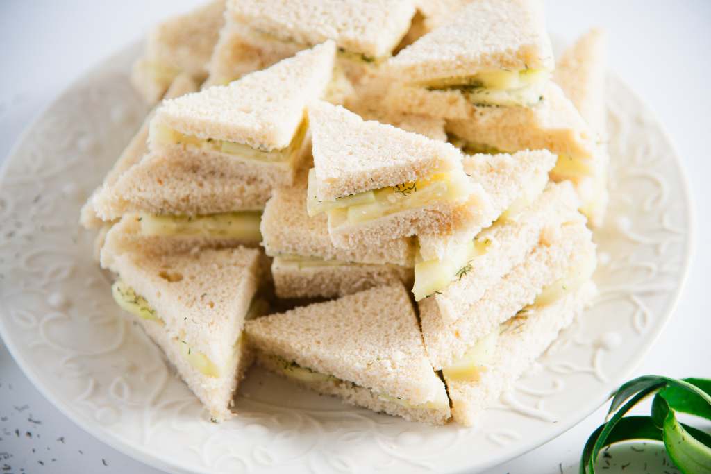 cucumber sandwiches stacked up on a plate, there is some spilled dill weed to one side of the plate, and some dill weed and cucumber peels on the other