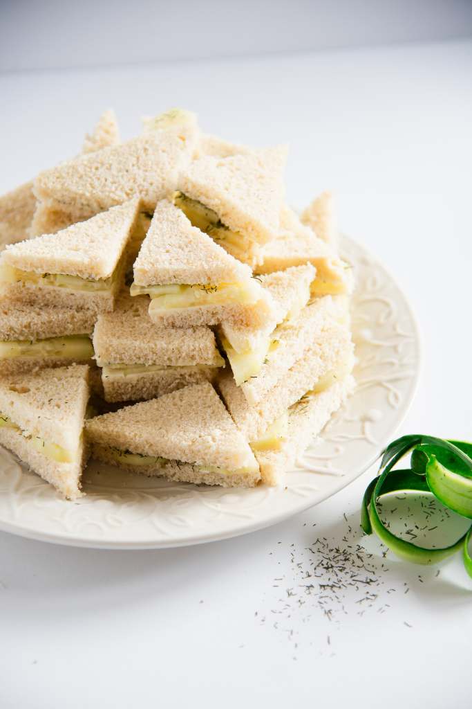 a bunch of sandwiches on a white plate, with some cucumber peels and spilled dill weed on one side of the plate