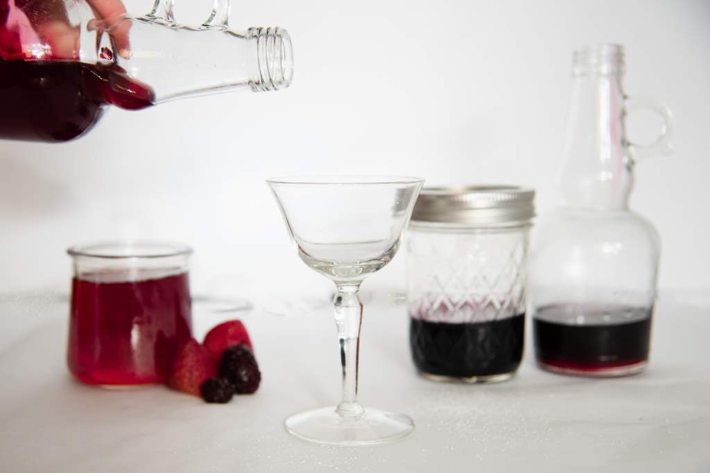 someone about to pour syrup into a glass cup. There are other bottles and jars with berry syrup in it