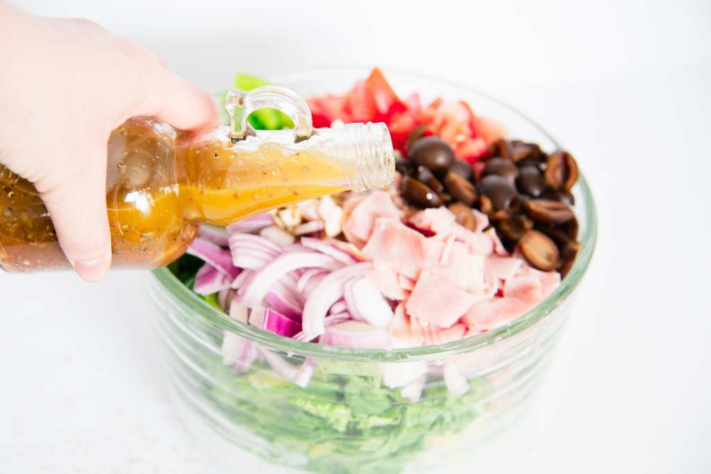 someone holding a bottle of salad dressing a above a bowl of salad about to pour it on