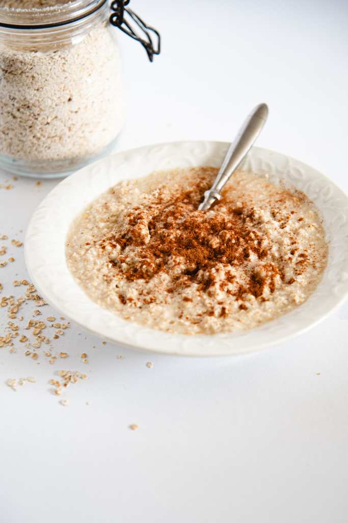 a bowl of cooked oatmeal with a spoon in it and cinnamon on top of it, there are some spilled oats by the bowl and the glass container of oatmeal