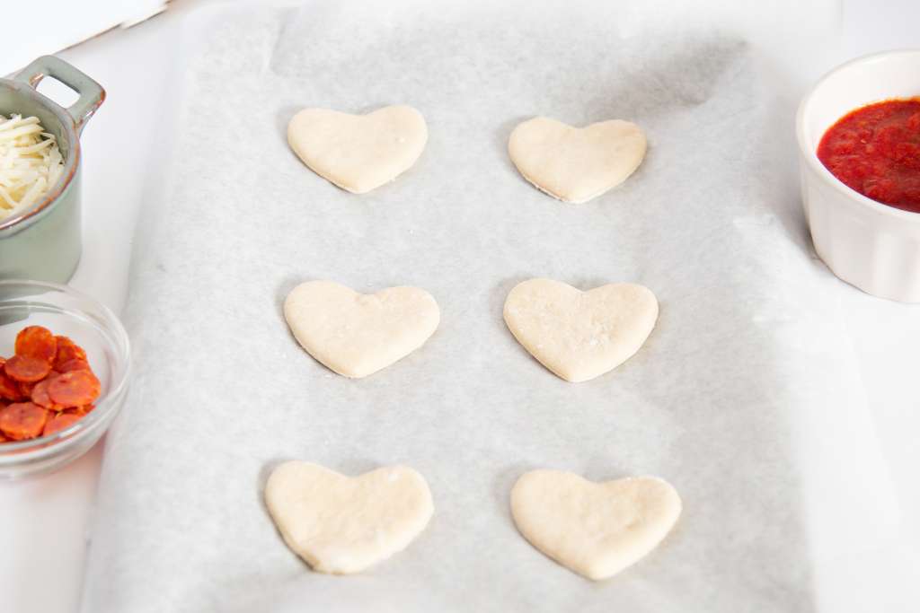 mini hearts cut out of pizza dough, sitting on a baking sheet, there are some pizza toppings next to the sheet