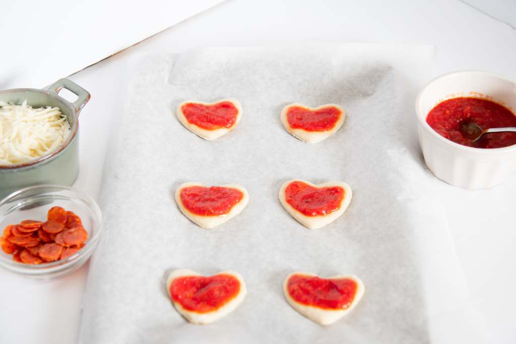 uncooked mini heart shaped pizzas on a baking sheet with other pizza toppings next to them