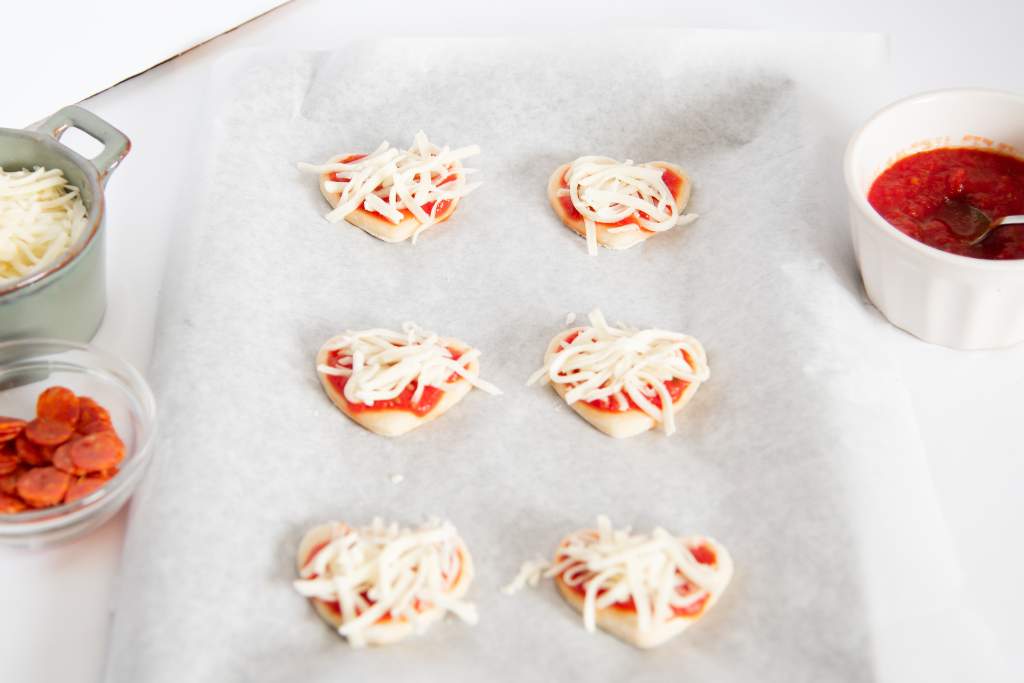 uncooked heart pizzas with sauce and shredded cheese on top, there are other pizza toppings next to them 