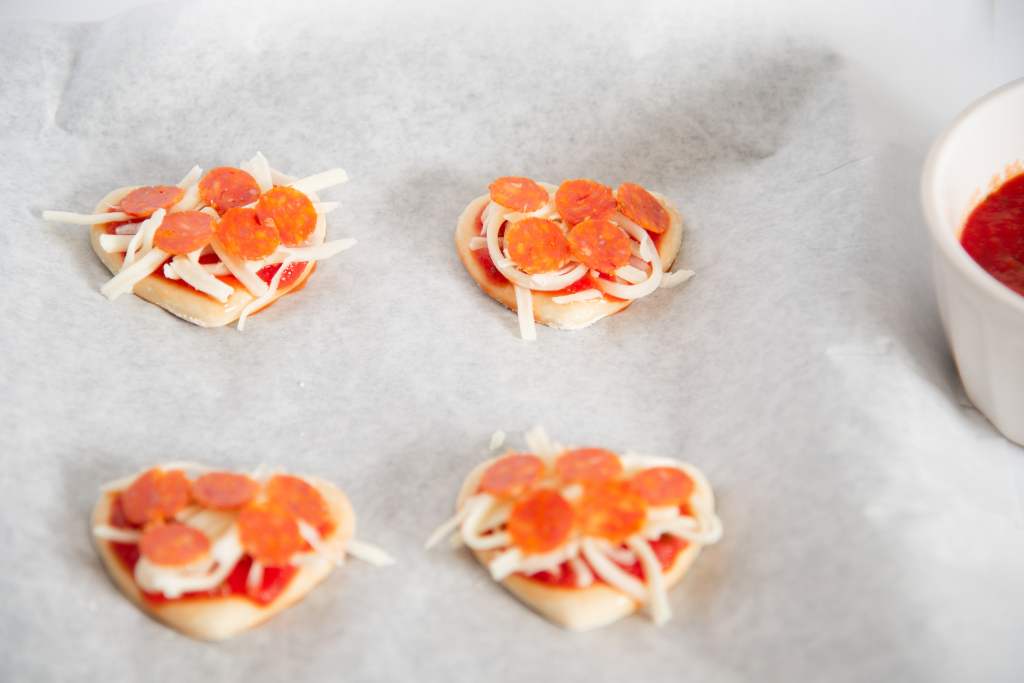 a close up of uncooked mini heart shaped pizzas with sauce, cheese, and pepperonis on top. You can see part of a dish of sauce next to them