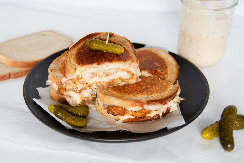 two cut sandwiches on a plate, one half on sandwich is sitting on top of there others, there are some pickles, some bread slices and a jar of dressing by the plate