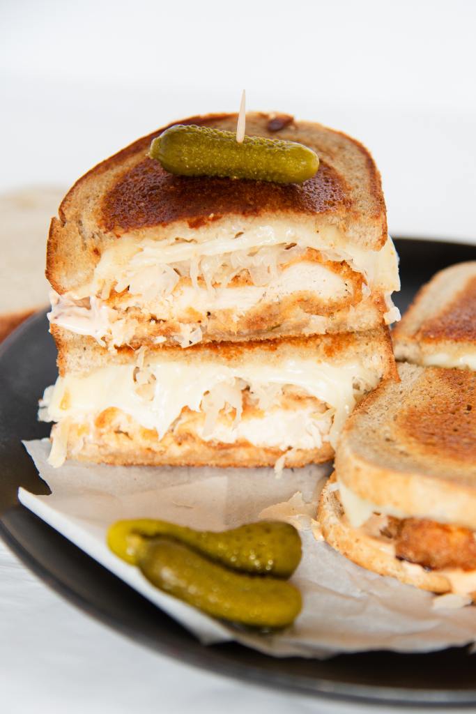 two halves of a chicken reuben stacked up top of each other, the top one has a pickle stuck in it, there are a few other pickles and another sandwich on the plate