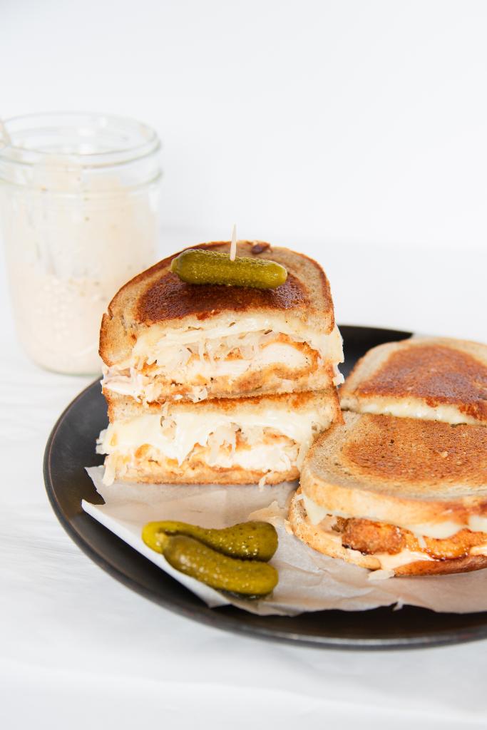 two sandwiches cut in half on a plate, two halves are stacked on top of each other, and the top on has a pickle stuck in it. There are a couple pickles on the plate and jar of dressing behind the plate