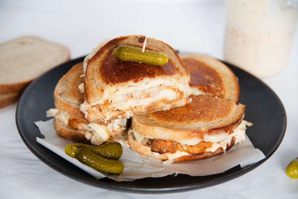 four chicken reuben sandwich halves on a plate, one is stacked on top of the others and has a pickle stuck in the top of it