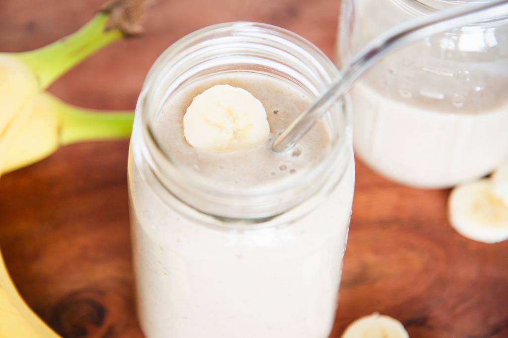 an overhead shot of a banana shake in a banana slice on it and a metal straw in it. You can see part of another shake and some bananas next to it 