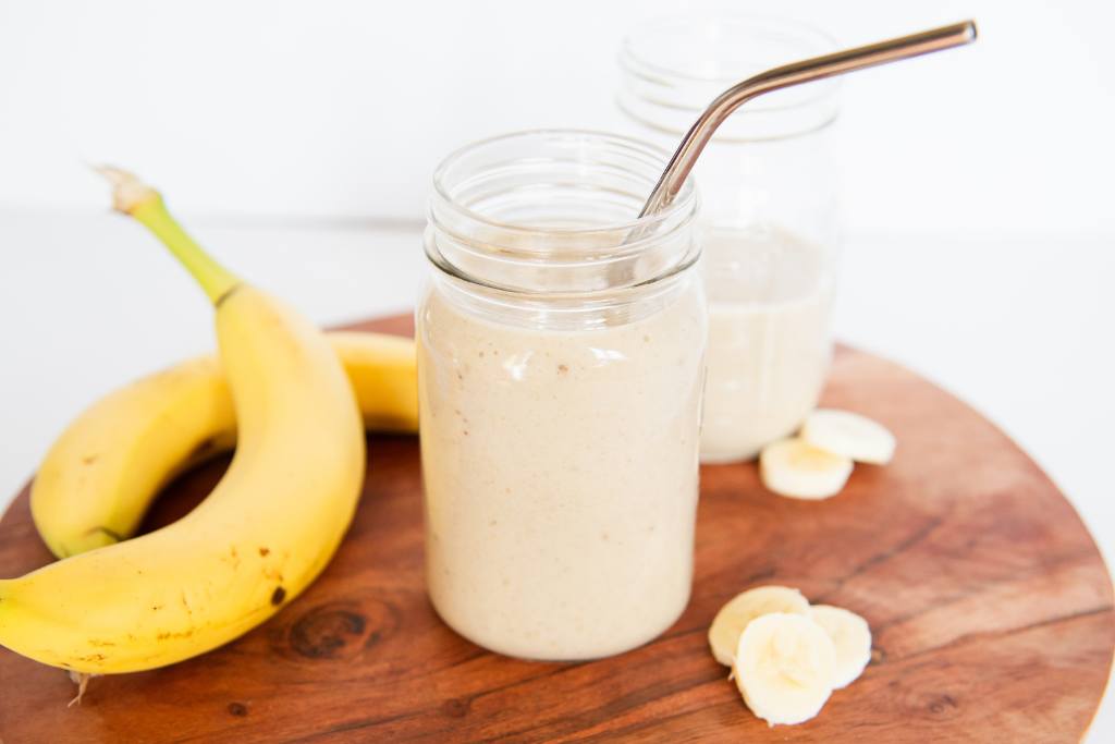 a banana shake with a straw in it on a wooden board, there is another shake behind it, and some whole bananas and banana slices next to it