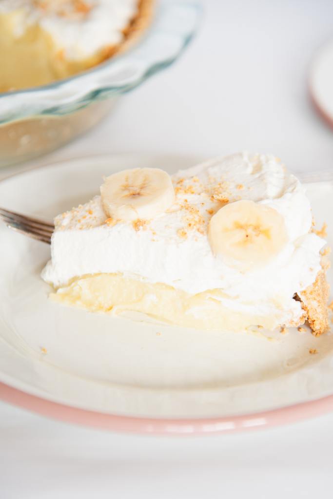 a slice on banana cream pie on a plate with a fork, you can see the rest of the pie next to it