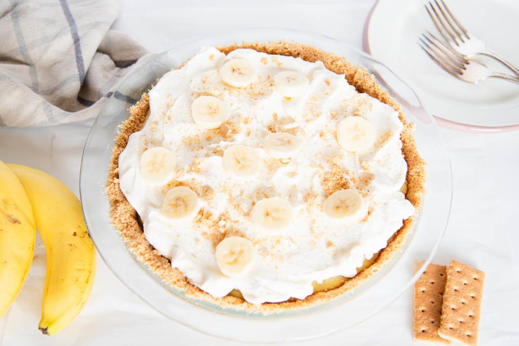 a finished banana cream pie with some plates, forks, whole bananas, graham crackers and a tea towel by it 