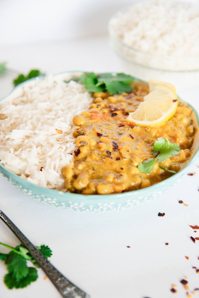 a bowl of curry and rice with lemon slices and cilantro on top, there is a fork in front of the bowl and a bowl of rice behind it