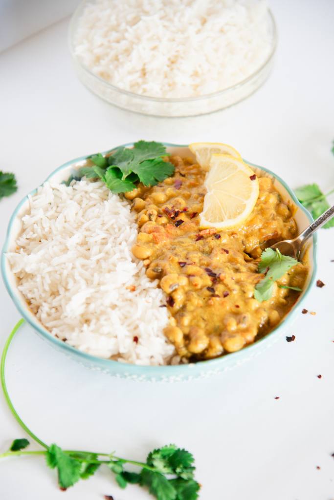 a bowl of curried black-eyed peas and rice with a fork in it, and a some lemon slices and cilantro leaves on it, there is also a bowl of rice behind it