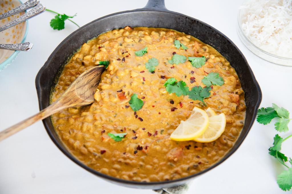 a pan of curry with a wooden spoon in it, there are some lemon sliced and cilantro leaves on top