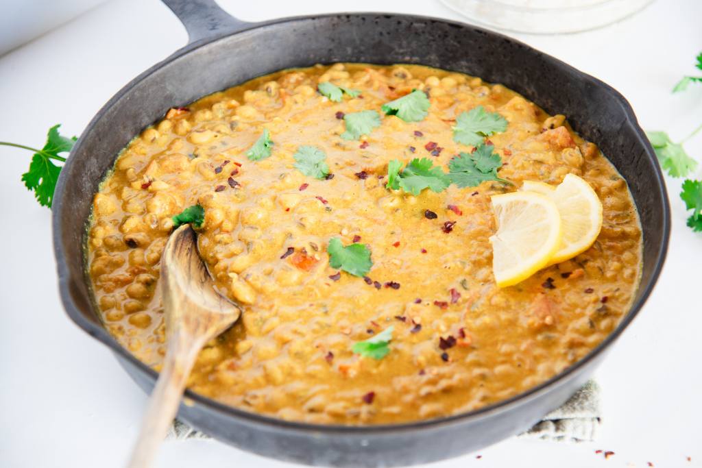 a pan of curried black-eyed peas with a wooded spoon in it, there are some lemon slices and cilantro leaves on top, and some cilantro next to the pan