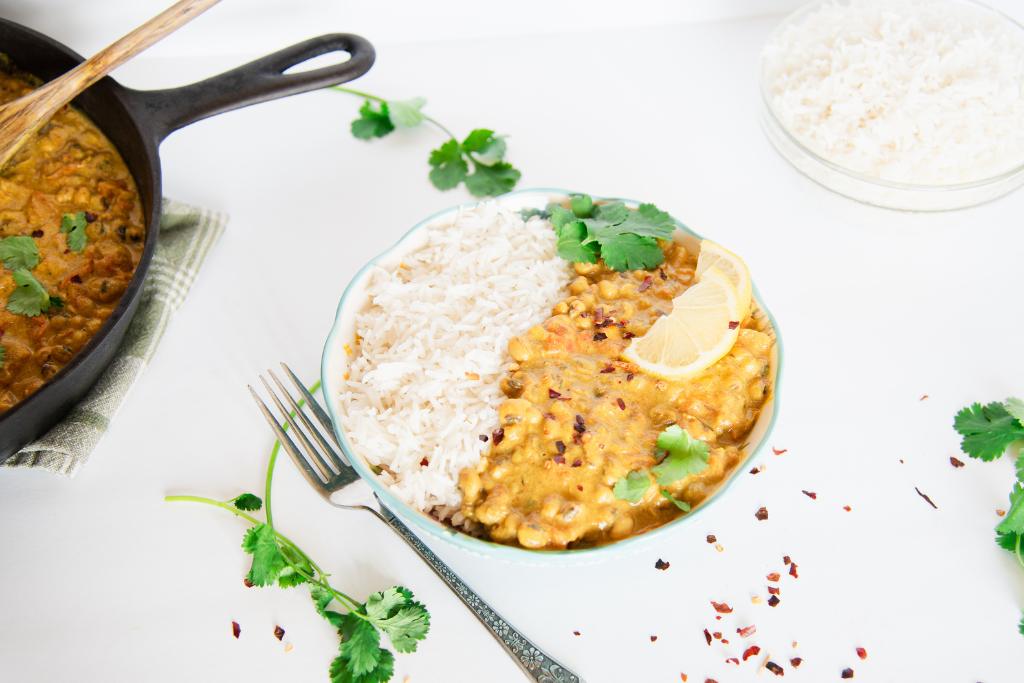 a bowl of curry and rice with lemon slices and cilantro leaves on top, there is a fork and some cilantro springs next to the bowl as well as a bowl of rice and a pan of curry 