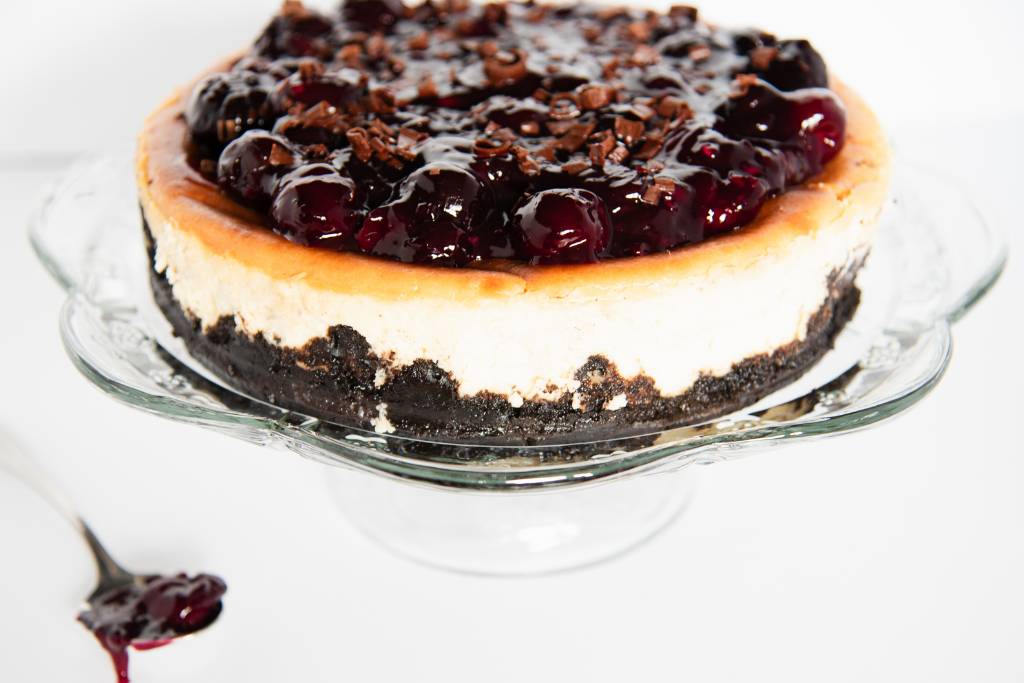 a cheesecake on a glass cake stand with a spoon with cherries next to it