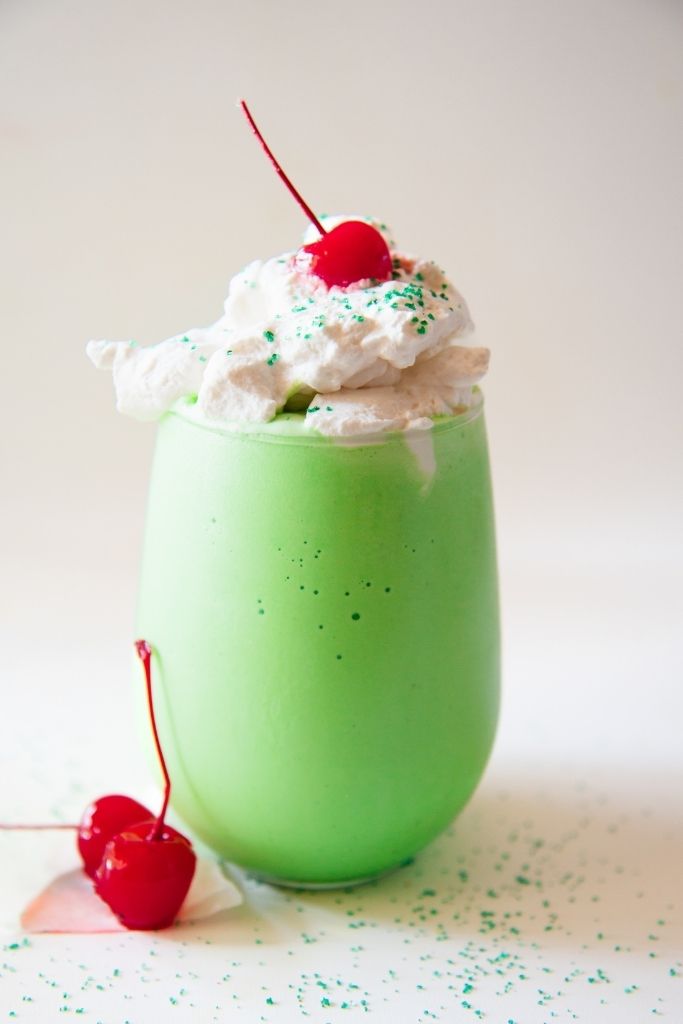 a Shamrock shake topped with whipped cream and a cherry, there are also some cherries next to the glass