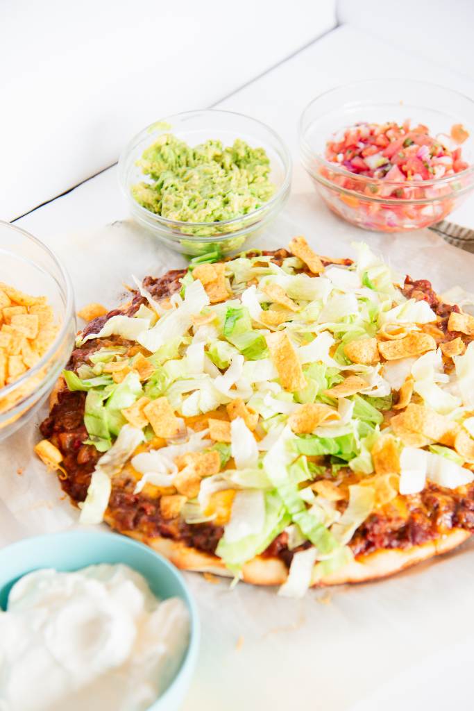 a pizza with just lettuce and chips on it, there are toppings in bowls around it