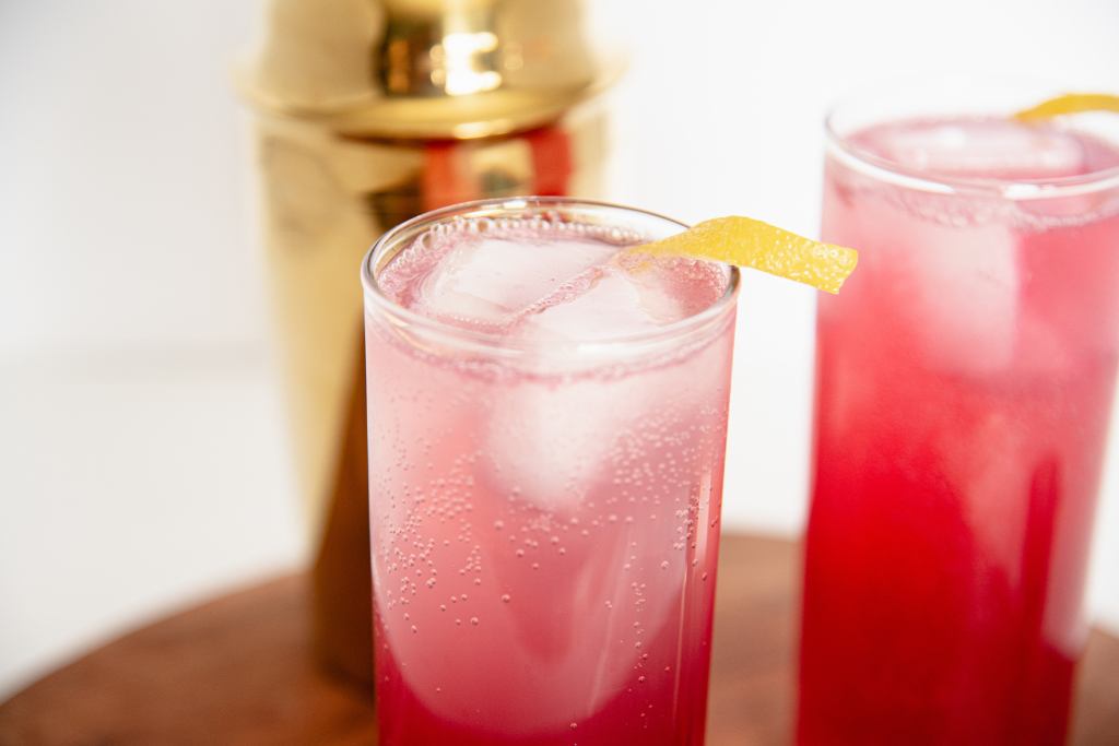 a close up of the top of a hard seltzer with two ice cubes in it and a lemon twist on top, there is another seltzer to the right side of it, and a gold cocktail shaker behind it